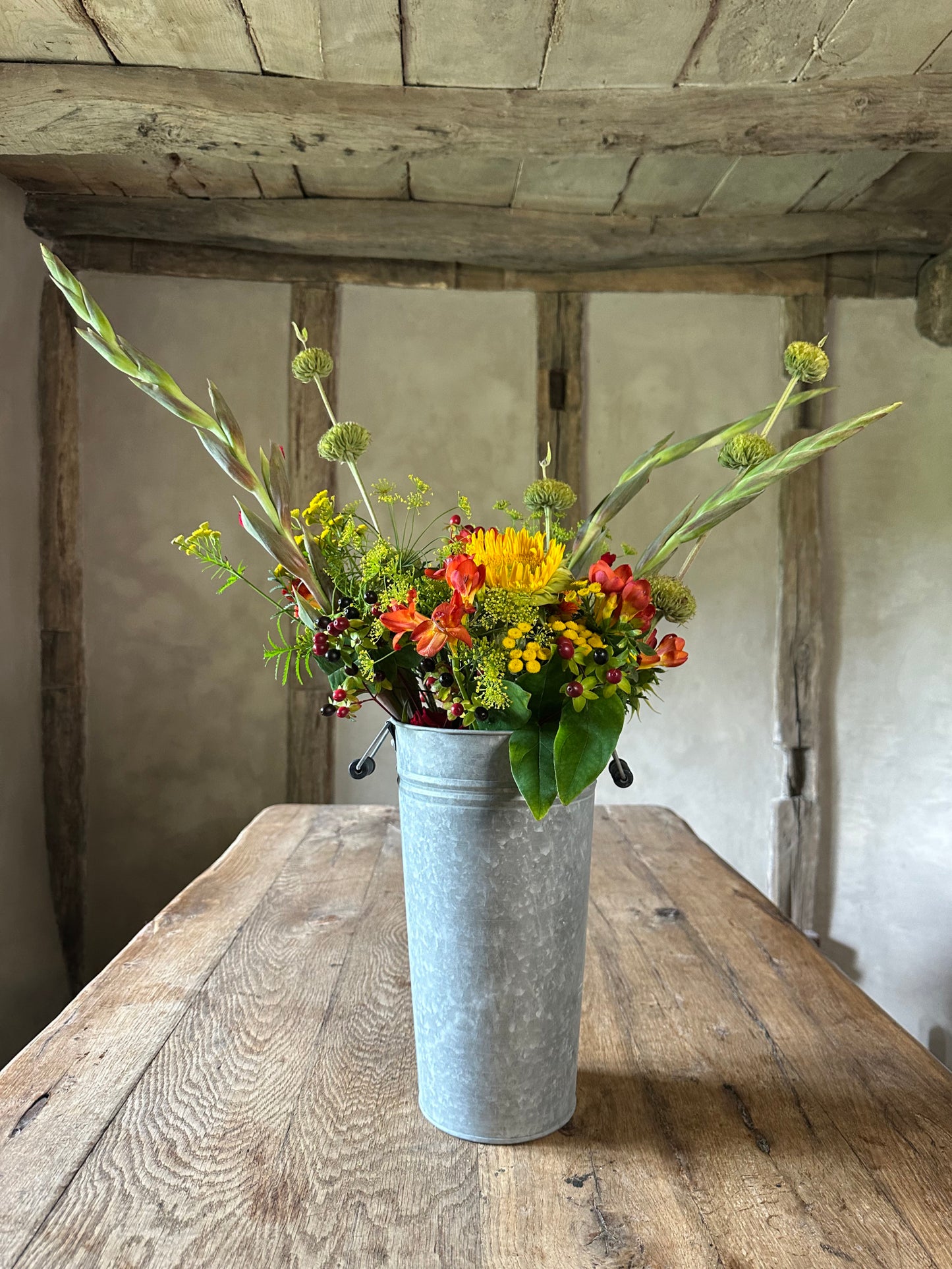 Floral arrangement in a metal bucket on a wooden table with rustic wooden beams above.