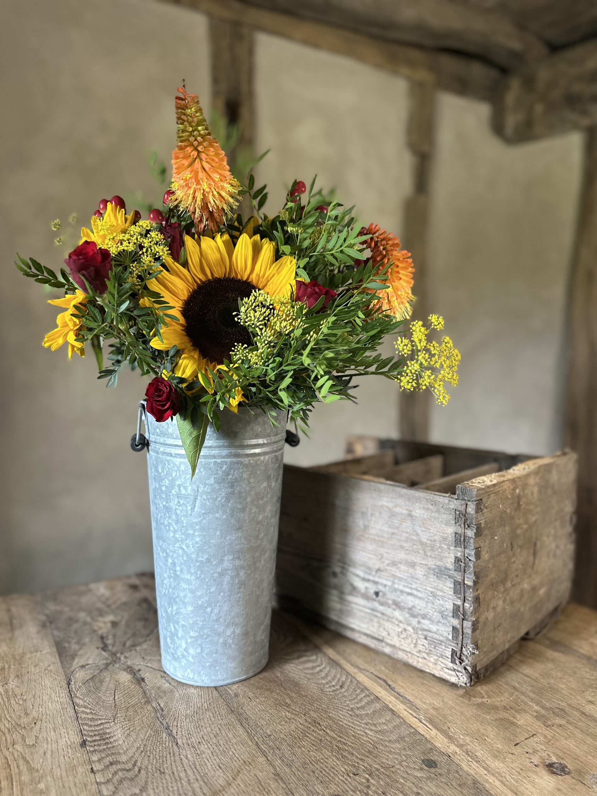 Vase with sunflowers and other flowers next to a wooden crate on a wooden floor.
